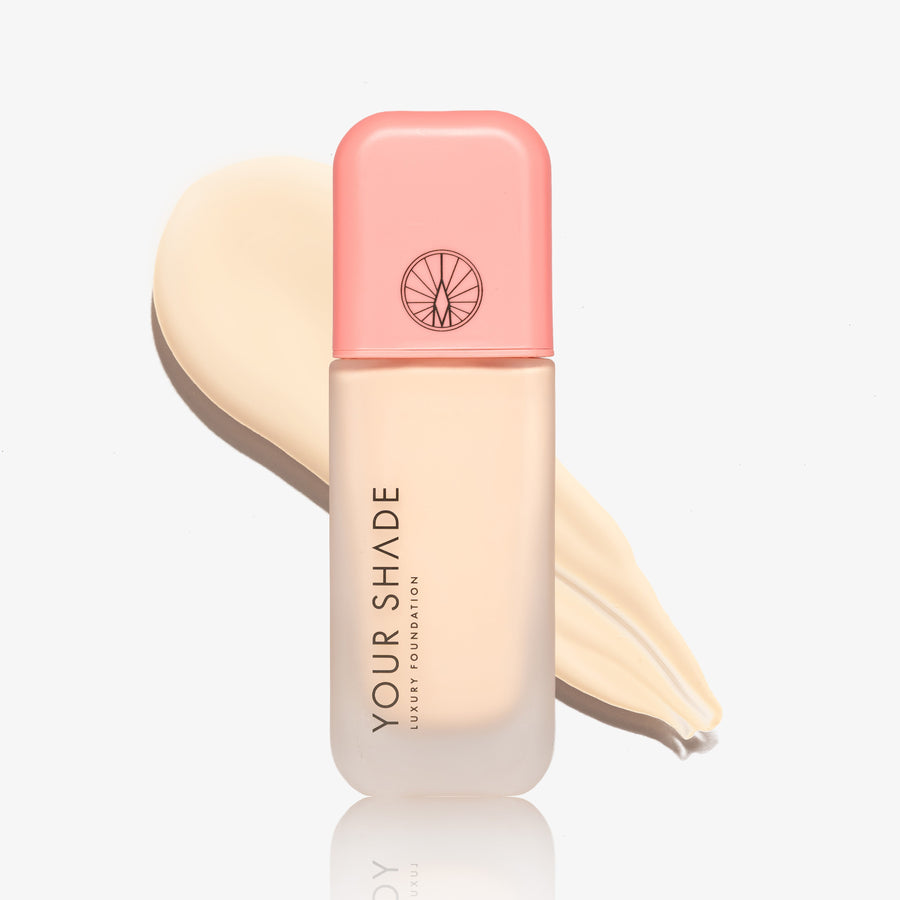 A bottle of Your Shade Foundation with a pink cap and frosted bottle showing the beige foundation inside, set against a cream-colored makeup smear on a white background.