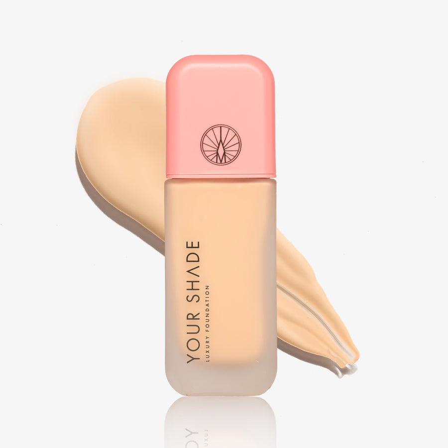 A bottle of luxury foundation by Your Shade, featuring a peach-pink cap and a frosted glass bottle showing the beige foundation liquid inside, set against a matching beige liquid swatch on a clean white background.