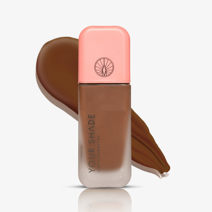 A bottle of liquid foundation with a peach-pink cap stands in front of a brown makeup swatch on a clean white background. The bottle is labeled 'YOUR SHADE'.