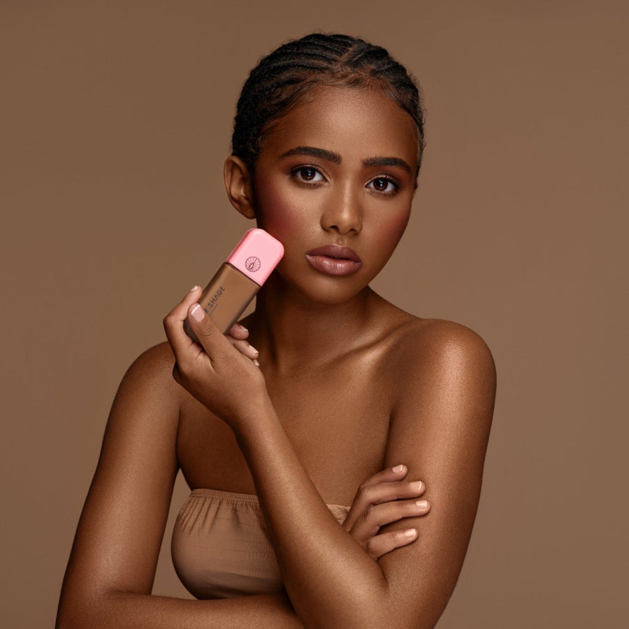 A woman with braided hair and warm medium-dark skin holds a pink-capped bottle of liquid foundation against her cheek, posed against a solid warm brown studio background.