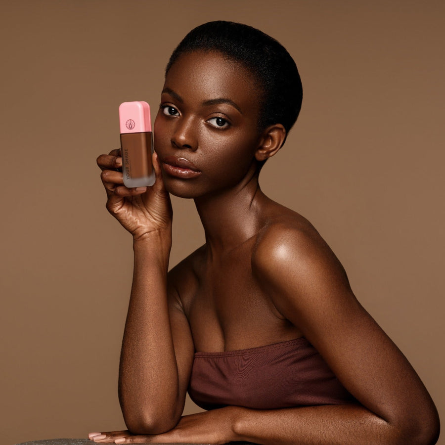 A woman with short dark hair holds up a bottle of liquid foundation with a pink cap against a solid warm brown studio background.