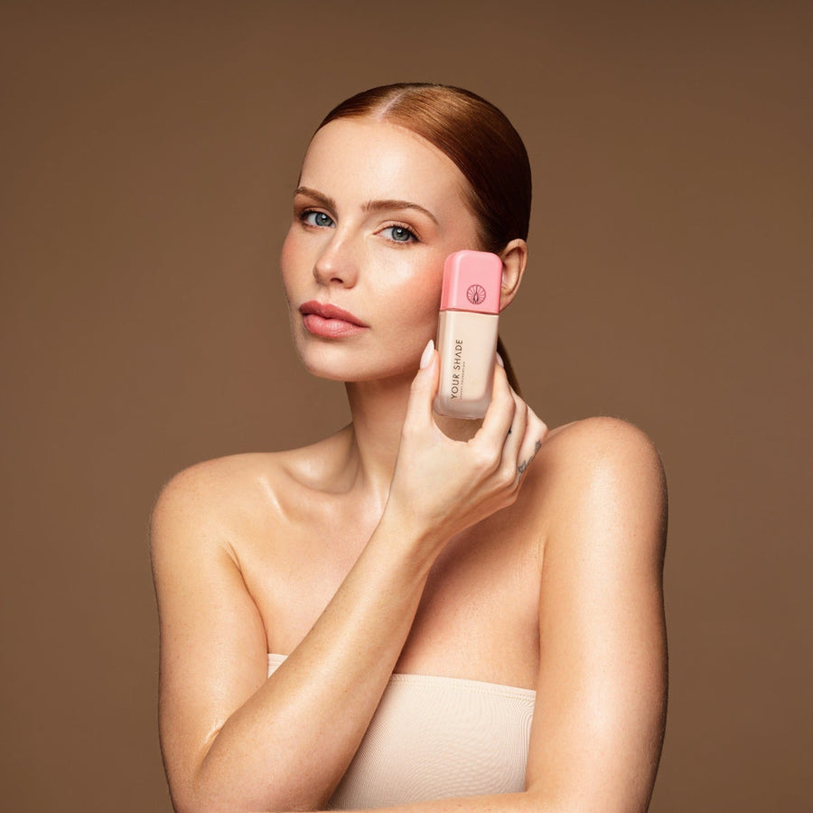 A woman with her hair pulled back holds a pink and beige cosmetic bottle against her cheek, set against a solid warm brown studio background.