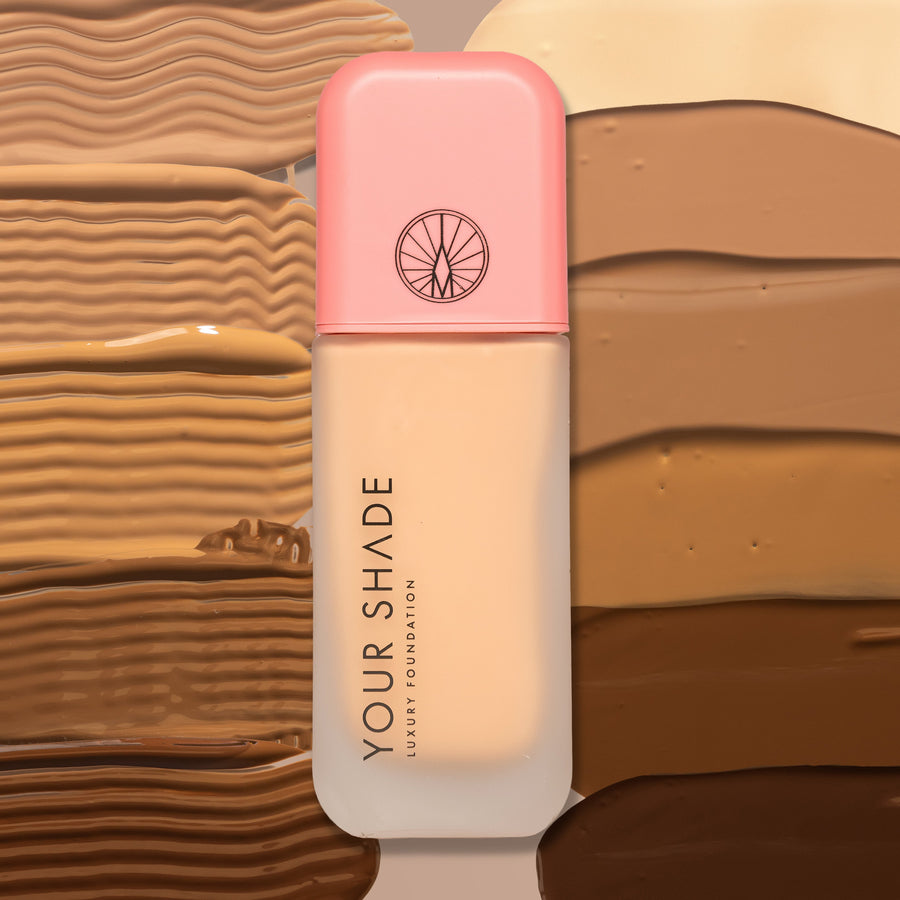 A bottle of Your Shade Luxury Foundation with a pink cap and frosted bottle showing the beige foundation inside, set against a background of textured swatches of various foundation shades.